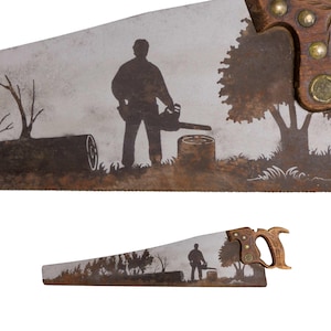 Könnte beinhalten: Eine Handsäge mit einer bemalten Szene auf dem Metallblatt. Das Kunstwerk zeigt die Silhouette einer Person mit einer Kettensäge, die neben einem Baumstumpf, einem gefällten Baumstamm und einem Baum steht. Der Griff ist aus Holz und mit Messingnieten befestigt.