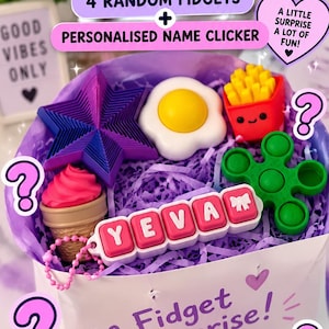 May include: A white gift box filled with purple paper shreds containing a mystery fidget pack. Items include a purple star, a fried egg, a red french fry container, a pink ice cream cone, a green pop-it, and a personalised clicker reading YEVA.