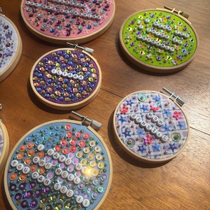 May include: Five circular wooden embroidery hoops containing fabric backgrounds in pink, lime green, purple, light blue, and a plaid pattern. Each hoop is decorated with colorful sequins, beads, and white letter beads forming phrases like "OH MY GOD HOLLANDER YOU ARE SO BORING," "I MEAN YES I THINK SO PROBABLY," "ILYAAA," "WOW GENETIC," and "I HAVE MY OWN WELL YOU CAN SEE IT."