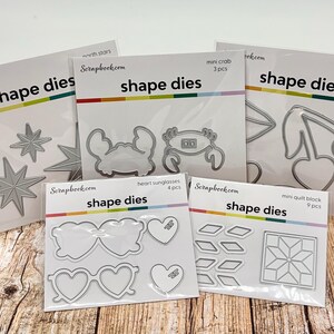 May include: Five packages of silver metal shape dies from Scrapbook.com. The sets include designs for north stars, a mini crab, cherries, heart sunglasses, and a mini quilt block. Each clear plastic package displays the die shapes inside.