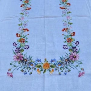 May include: A rectangular white tablecloth featuring a central embroidered floral border. The design consists of colorful cross-stitched flowers in shades of pink, orange, yellow, blue, and purple, arranged in a U-shaped pattern across the fabric.