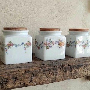 Puede incluir: Tres botes cuadrados de cerámica blanca con esquinas redondeadas y tapas de madera planas. Cada bote presenta un delicado patrón de guirnalda floral en tonos rosa, azul, amarillo y verde alrededor del cuerpo de porcelana blanca.