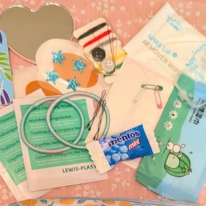 May include: A collection of personal care and emergency items including a heart-shaped mirror, a patterned nail file, sewing kit, safety pins, hair pins, Mentos mints, wound cleansing wipes, makeup remover wipes, and a small blue packet with text.