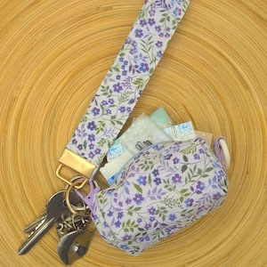 May include: A fabric wristlet keychain featuring a white background with a purple and green floral pattern, attached to a gold-tone metal clasp. A matching small zippered pouch with the same floral print is connected to the keychain.