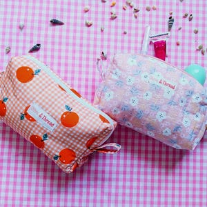 May include: Two rectangular fabric pouches with zip closures. One pouch features an orange and white gingham pattern with orange fruit illustrations and a white label reading Olive & Thread. The other is pink with white flowers and a matching label.