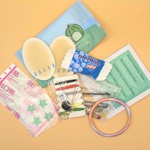 May include: A collection of small items including two oval adhesive bandages, a Mentos mint roll, a sewing kit with thread and buttons, a green wound cleansing wipe packet, a pack of star-patterned bandages, and a thin, circular, gradient-coloured bangle.