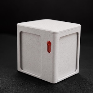May include: A square, off-white box with a speckled texture and a recessed panel design on the side. A small, vertical, translucent red rectangular indicator light is embedded into the front face of the box, which has a flat top surface.