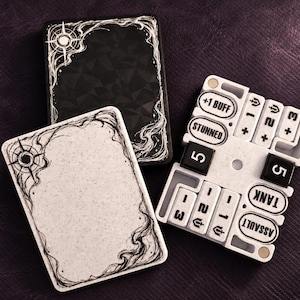 May include: Two rectangular game cards with black and white ornate borders and a white game tracker board featuring black and white tiles labeled with text including +1 BUFF, STUNNED, TANK, ASSAULT, and the number 5, arranged in a grid.