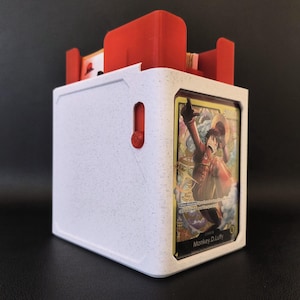May include: A white speckled plastic trading card deck box with a red interior mechanism and a red side button. The front of the box features a clear display window holding a Monkey D. Luffy trading card with visible text and character art.