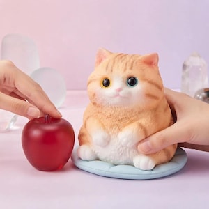 May include: A small, orange tabby cat figurine with white paws and chest, sitting on a light blue circular cushion. Beside the cat is a smooth, translucent red apple-shaped object. The cat has large, round eyes with distinct colors.