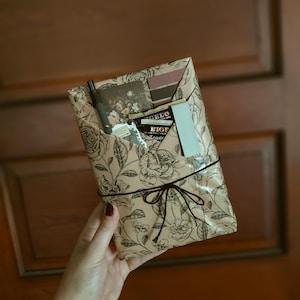 May include: A rectangular package wrapped in beige paper with a black floral line art pattern, secured with dark brown twine tied in a bow. The package contains a dark pen, a small floral card, a Bigelow tea bag, and a light blue rectangular tag.