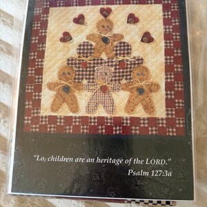 May include: A greeting card featuring a quilted design of five gingerbread figures in a pyramid arrangement, accented with small red hearts and buttons. The text reads, "Lo, children are an heritage of the LORD." Psalm 127:3a, set against a dark background.