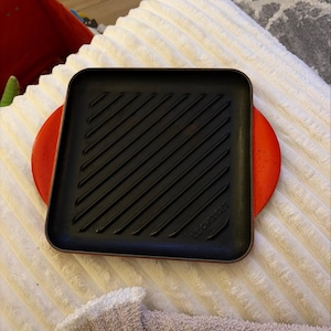May include: A square Le Creuset grill pan with a black interior featuring diagonal ridges and a vibrant orange exterior. The pan has rounded corners and integrated side handles, with the brand name embossed clearly on the bottom right corner.