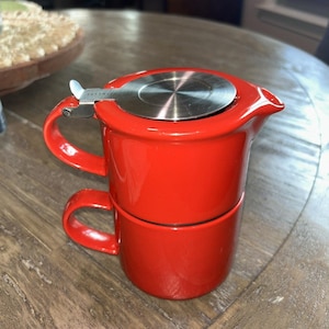 May include: A bright red ceramic tea set consisting of a teapot stacked directly on top of a matching mug. The teapot features a stainless steel lid with a small metal lever handle and a spout, while both pieces have integrated side handles.