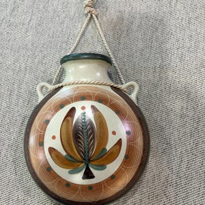 May include: A round ceramic wall vase with a cream-colored center featuring a painted brown and gold floral motif. The vase has a dark green rim, two side handles, and a beige rope hanger attached to the top for mounting on a vertical surface.