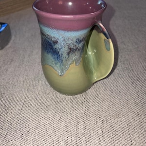 May include: A ceramic mug featuring a smooth, rounded body with a unique integrated handle design. The exterior displays a multi-colored glaze transition, starting with a deep plum rim, blending into a vibrant blue midsection, and ending in a light olive green base.