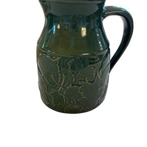 May include: A dark green ceramic pitcher featuring a glossy finish and a relief pattern of maple leaves around the lower body. The vessel has a curved handle on the right side, a wide cylindrical neck, and a prominent pouring spout at the top rim.