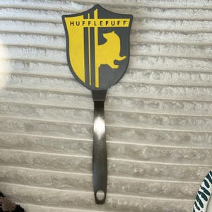 May include: A kitchen spatula featuring a shield-shaped head with a dark gray background, a yellow badger silhouette, and three vertical yellow stripes. The word HUFFLEPUFF is printed in black text at the top. It has a long, silver metal handle.