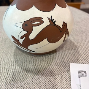May include: A round, hand-painted ceramic pot featuring a cream-colored body with a brown rim and base. The exterior is decorated with a brown graphic illustration of a leaping deer, outlined in black, set against the cream background.