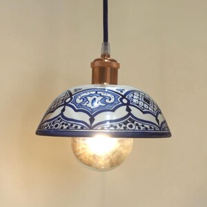 May include: A dome-shaped pendant light featuring a white ceramic shade with intricate blue hand-painted patterns. The fixture includes a metallic copper-coloured socket, a black cord, and a clear glass light bulb emitting a warm glow.