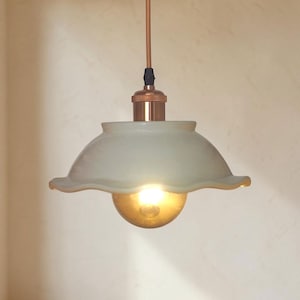 Könnte beinhalten: Eine Pendelleuchte mit einem glänzenden, cremefarbenen Keramikschirm mit gewelltem, gerüschtem Rand. Die Leuchte verfügt über eine kupferfarbene Fassung und ein passendes Kabel sowie eine warm leuchtende, bernsteinfarbene Glühbirne.
