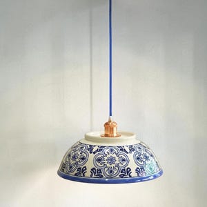 May include: A pendant light featuring a ceramic bowl shade with a blue and white floral pattern. The fixture includes a copper-coloured ceiling canopy and socket, connected by a blue cord. The text InutileStudio is visible in the bottom right.
