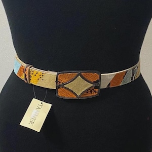 May include: A belt featuring a multi-colored snakeskin pattern in shades of orange, yellow, and grey. The rectangular buckle has a central diamond shape with a tan inlay. A rectangular tag with the text TANNER is attached to the belt strap.