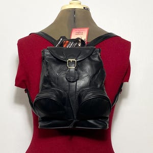 May include: A black leather backpack featuring a flap closure with a gold-tone metal buckle, two front zippered pockets, and adjustable shoulder straps. The bag includes a red tag with the text Genuine Leather and a black tag with white text.