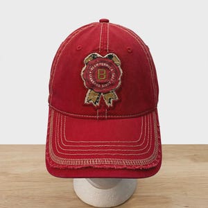 May include: A red baseball cap with a distressed, frayed brim and light-colored contrast stitching. Centered on the front is a circular patch featuring a gold letter B, the text BEAM FORMULA and A STANDARD SINCE 1795, shaped like a wax seal.