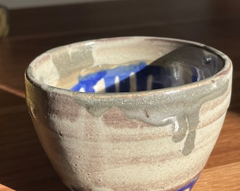 May include: A small, handmade ceramic bowl with a light brown glaze and blue accents. The bowl has a slightly irregular shape and a textured surface.
