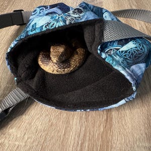 May include: A blue fabric pouch with a Celtic knot pattern and a grey adjustable strap, featuring a soft black fleece lining. A brown and tan patterned snake is curled inside the open bag, resting on the dark interior fabric surface.