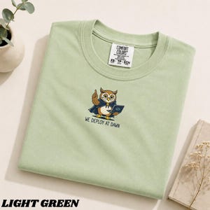 Puede incluir: Una camiseta de color verde claro con un gráfico de un búho con una capa azul oscuro, sosteniendo un portátil y señalando hacia arriba. Debajo del búho, el texto dice "WE DEPLOY AT DAWN". La camiseta tiene una etiqueta Comfort Colors en el cuello.