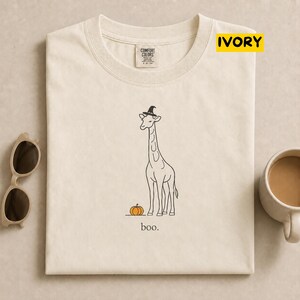 May include: An ivory t-shirt featuring a black line art illustration of a giraffe wearing a witch hat standing next to an orange pumpkin. Below the giraffe, the word "boo." is printed in a simple black font. The shirt has a Comfort Colors label.