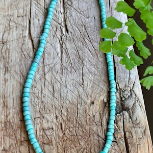 May include: A beaded necklace featuring small, matte turquoise-coloured round beads strung in a continuous loop. The necklace includes a single light pink bead accented by a small, silver-toned spacer bead with embedded blue crystals at the centre.