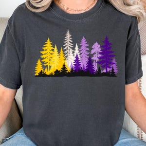 May include: A charcoal grey short-sleeve t-shirt featuring a graphic print of a forest landscape. The trees are rendered in a gradient of colours including yellow, white, and various shades of purple, set against a solid black ground line.