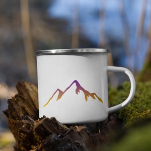 May include: A white enamel mug featuring a gradient mountain range graphic that transitions from purple at the peak to orange and yellow at the base. The mug has a smooth white finish and a silver-colored metal rim around the top edge.