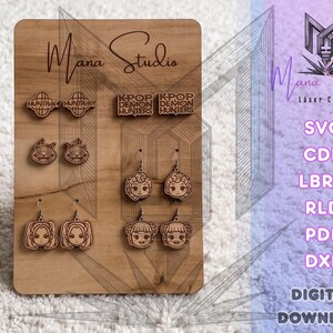 Puede incluir: Un tablero de exhibición de madera marrón claro con pendientes cortados a láser con varios diseños, incluyendo logotipos de globos, caras de gato y personajes de estilo anime. El tablero está grabado con el texto Mana Studio y KPOP DEMON HUNTERS.