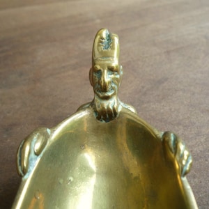 May include: A brass trinket dish shaped like a person with a beard and a hat. The dish is oval and has a smooth, shiny surface.