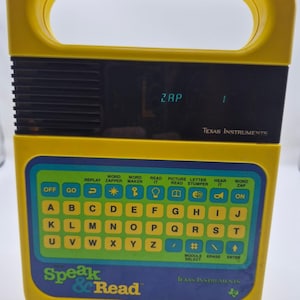 May include: A yellow and blue Texas Instruments Speak & Read electronic learning toy with a handle, a black speaker grille, and a digital display showing the text ZAP 1. The device features a QWERTY-style keyboard with various function icons.