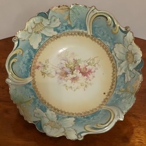 May include: A round porcelain bowl featuring a scalloped edge with embossed floral relief details. The rim is painted in shades of blue and cream, while the center displays a floral bouquet surrounded by a decorative gold filigree border.