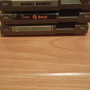May include: A stack of three gray Nintendo Entertainment System game cartridges. The top cartridge is labeled Marble Madness, the middle cartridge is labeled Ultra Q-bert, and the bottom cartridge is labeled Alpha Mission. Each cartridge has a rectangular shape.