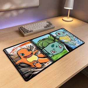 May include: A rectangular desk mat featuring a three-panel graphic illustration of three cartoon creatures. The left panel shows an orange fire-type creature, the centre panel displays a green plant-type creature, and the right panel depicts a blue water-type creature.