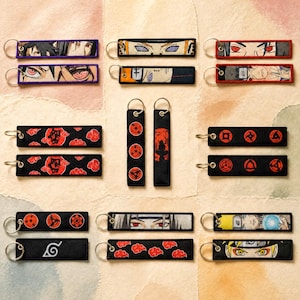 Könnte beinhalten: Eine Sammlung rechteckiger Schlüsselanhänger aus Stoff mit verschiedenen Anime-inspirierten Motiven, darunter Charakteraugen, rote Wolkenmuster und kreisförmige Symbole, jeweils befestigt an einem silberfarbenen Metallring.