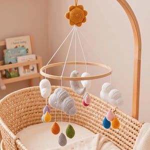 May include: A crochet baby mobile featuring a central yellow sun, a grey crescent moon, and several white and grey clouds. Multi-coloured crochet raindrops in shades of blue, pink, yellow, green, and purple hang from the clouds on white strings.