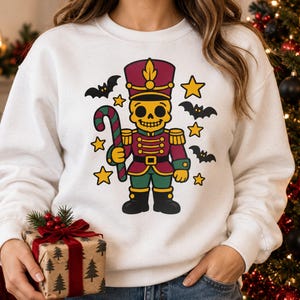 May include: A white crewneck sweatshirt featuring a graphic print of a yellow skull-faced nutcracker soldier wearing a maroon and green uniform, holding a candy cane, surrounded by black bats and yellow stars on the front center.