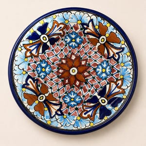 May include: A round ceramic plate featuring a hand-painted pattern with a central brown flower, surrounded by a red and green lattice design. The border includes alternating blue and white floral motifs, accented with dark blue and brown leaf shapes.