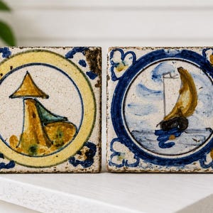 May include: Two square ceramic tiles with a distressed, crackled finish. The left tile features a yellow and blue circular border surrounding a stylized yellow structure. The right tile displays a blue circular border with a yellow sailboat illustration.