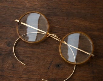 Könnte beinhalten: Runde Brille mit dünnen, bernsteinfarbenen kreisförmigen Rahmen und klaren Gläsern. Die Brille verfügt über einen goldfarbenen Metallsteg und schlanke, geschwungene Bügel, die nach außen ragen und ein klassisches, minimalistisches Design aufweisen.