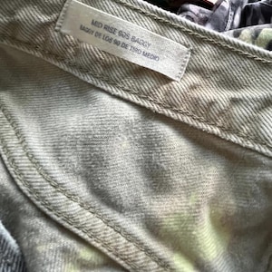 Pode incluir: Uma cal&ccedil;a jeans de cor clara com uma etiqueta vis&iacute;vel no c&oacute;s com o texto MID RISE 90S BAGGY e BAGGY DE LOS 90 DE TIRO MEDIO. O tecido apresenta um padr&atilde;o sutil e manchado em tons claros com toques de amarelo e cinza.