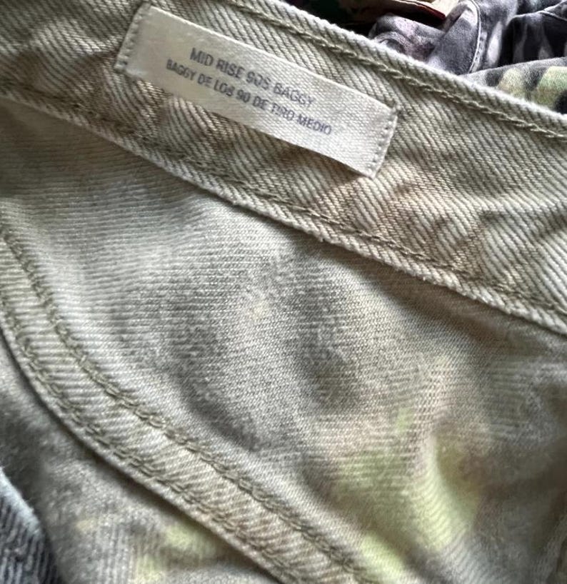 Pode incluir: Uma cal&ccedil;a jeans de cor clara com uma etiqueta vis&iacute;vel no c&oacute;s com o texto MID RISE 90S BAGGY e BAGGY DE LOS 90 DE TIRO MEDIO. O tecido apresenta um padr&atilde;o sutil e manchado em tons claros com toques de amarelo e cinza.